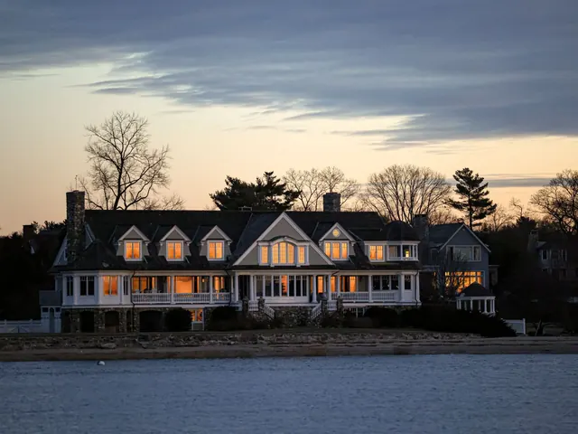 Large shingle-style waterfront estate at twilight with warm lit windows and a calm bay in front