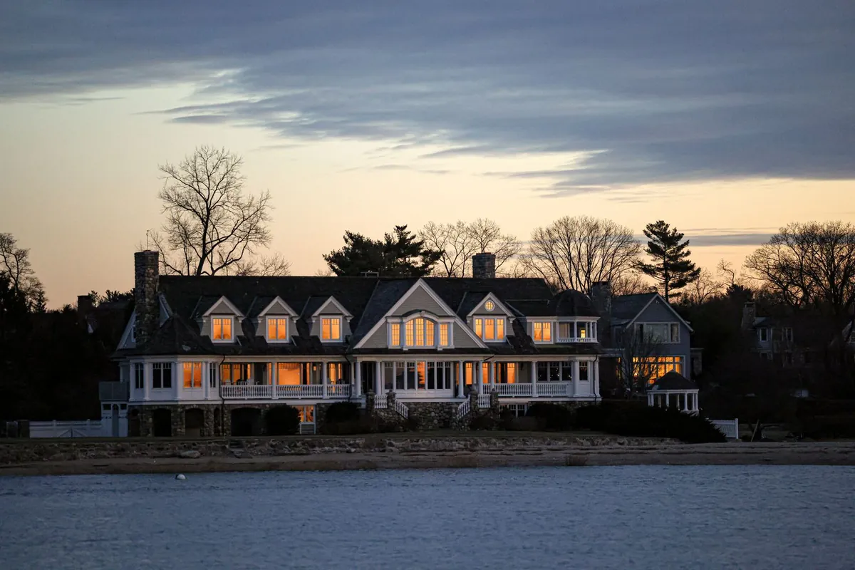 Large shingle-style waterfront estate at twilight with warm lit windows and a calm bay in front