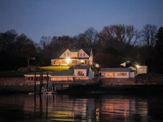 Lakeside house glowing at dusk with its lights reflected in dark calm water and a dock beside it