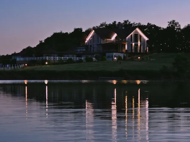 Lit house on a wooded shoreline at dusk with its lights reflected in a calm lake