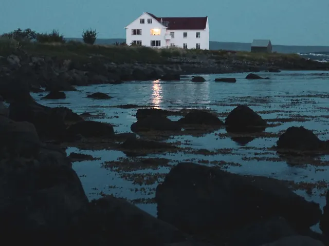 White house on a rocky shore at blue hour with its lights shining across dark water