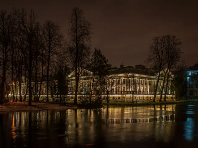 Large lit-up palace-style estate at night reflected in a still lake with bare trees in the foreground