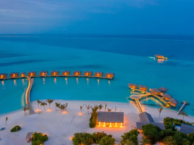 Aerial view of overwater villas with warm lights glowing at dusk on turquoise water