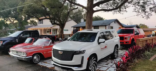 White Cadillac Escalade with a Red Star door sign parked beside a red F-150, a black F-250 and a red classic convertible at dusk
