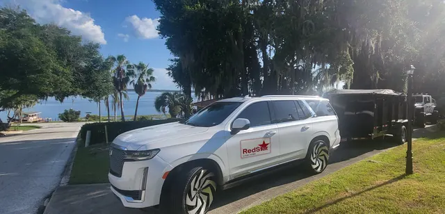 White Cadillac Escalade with a Red Star door sign and black and white wheels towing a black dump trailer beside a lake under oak trees