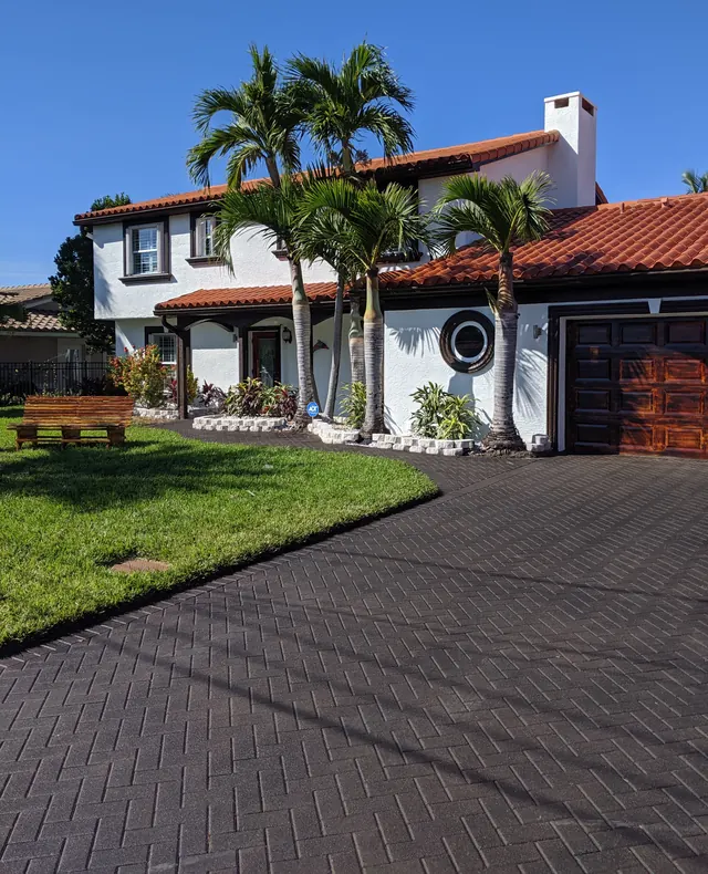 Herringbone paver drive leading to a white home with a red tile roof, palms, a porthole window and a wood garage door