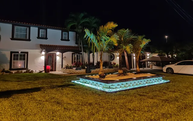 Angled night view of a white two-story home with uplit palms in a raised planter edged in blue-lit block