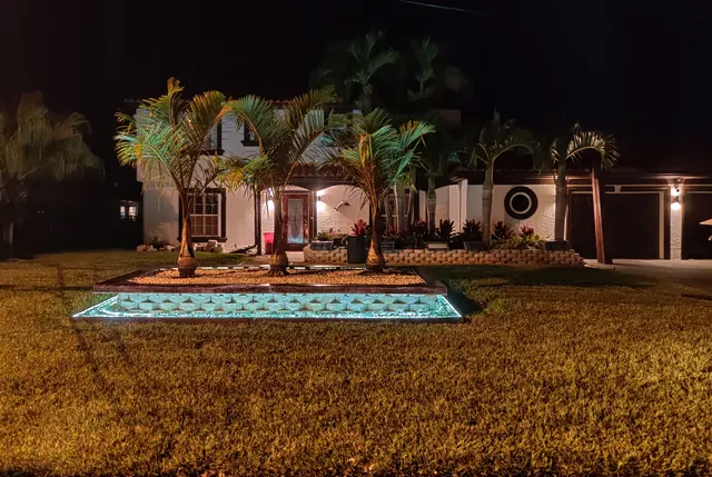 White two-story home at night with a raised palm planter edged in glowing blue-lit block on the front lawn