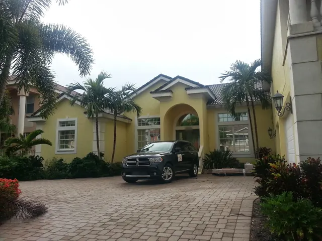 Yellow Mediterranean-style home with an arched entry, palms and a dark SUV parked on a paver drive