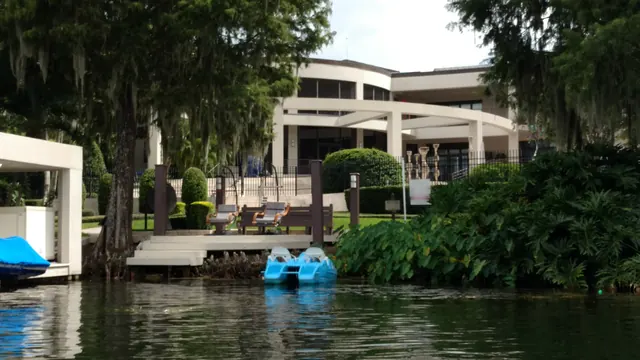 Lakefront home seen from the water, with a dock, lounge chairs, blue pedal boats and tropical plantings under moss-draped oaks