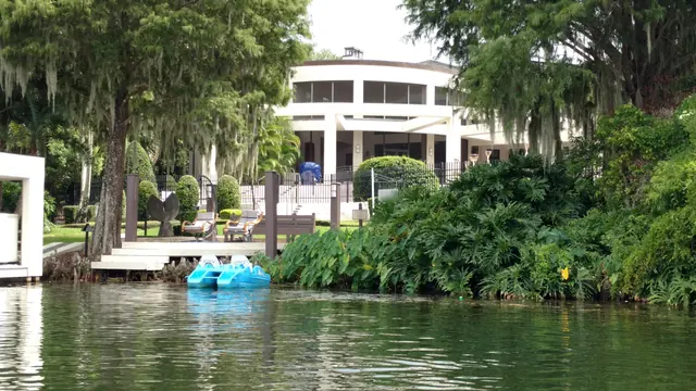 Wider view from the lake of a curved white lakefront home, its dock, blue pedal boats and lush shoreline plantings