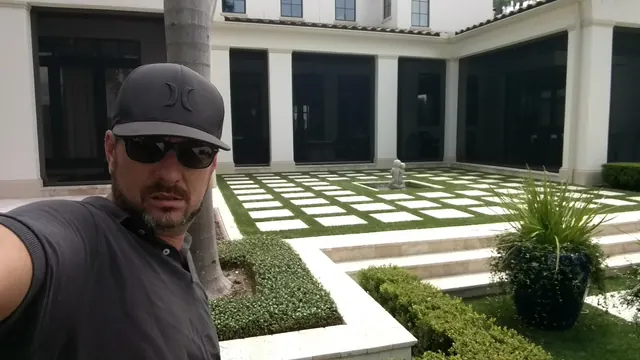 Man in a black cap and sunglasses taking a selfie in a courtyard of white pavers set in turf with boxwood hedges
