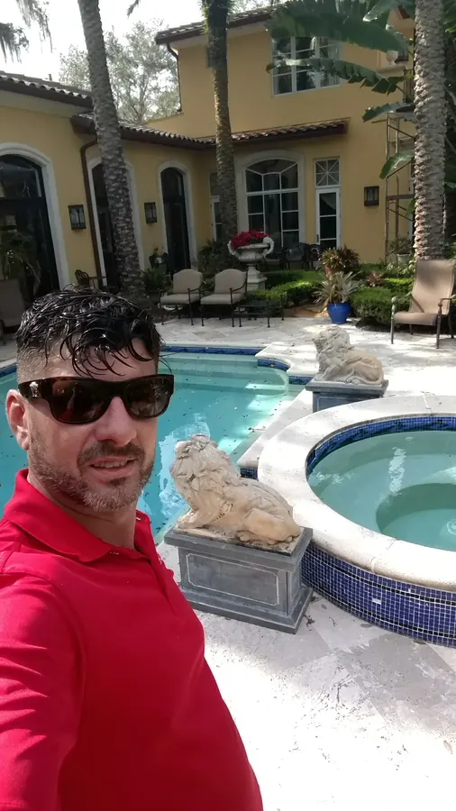 Man in sunglasses and a red shirt taking a selfie beside a pool and spa with stone lion statues at a Mediterranean-style home