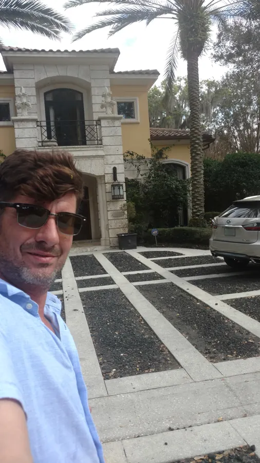 Man in sunglasses and a light blue shirt taking a selfie in front of a yellow Mediterranean estate with a paver-grid drive