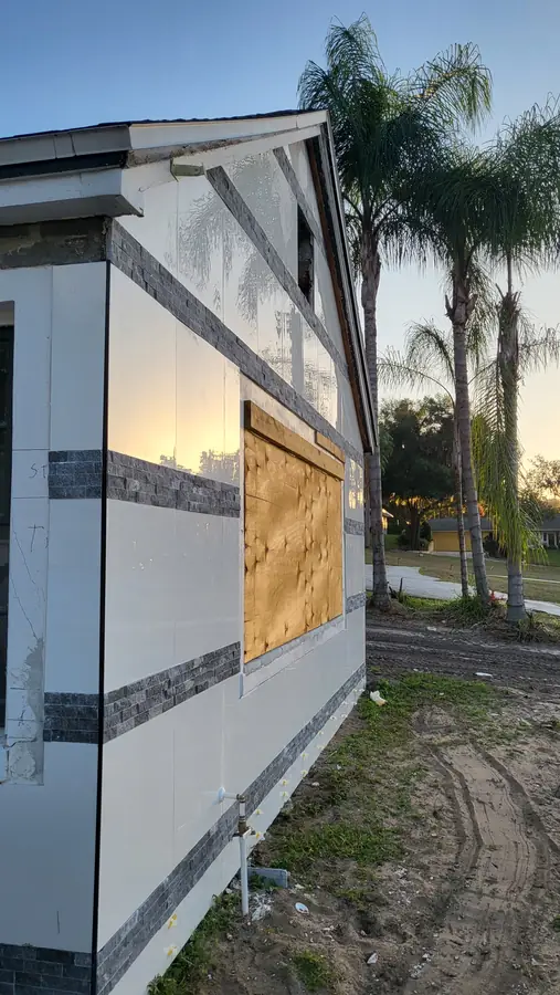 White exterior wall with dark stacked-stone accent bands at sunset, window still covered in plywood, palm trees behind