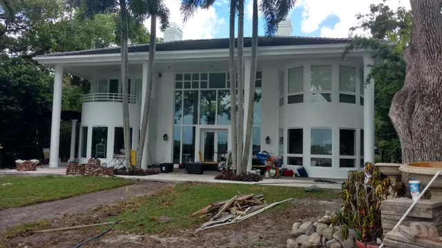 White two-story lakefront home with tall palms, pallets of materials and tools on the terrace and a regraded lawn in front